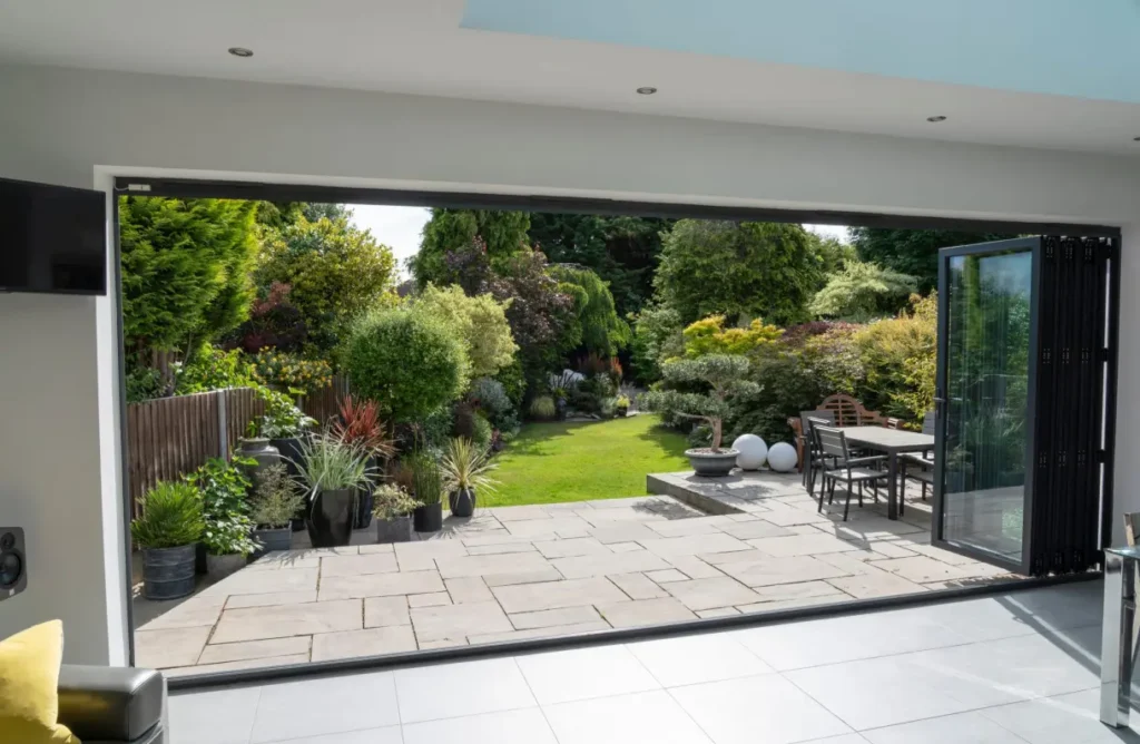 Aluminium bi-fold door system fully stacked to the side, creating a full-width, unobstructed opening between an indoor dining area and an outdoor garden space